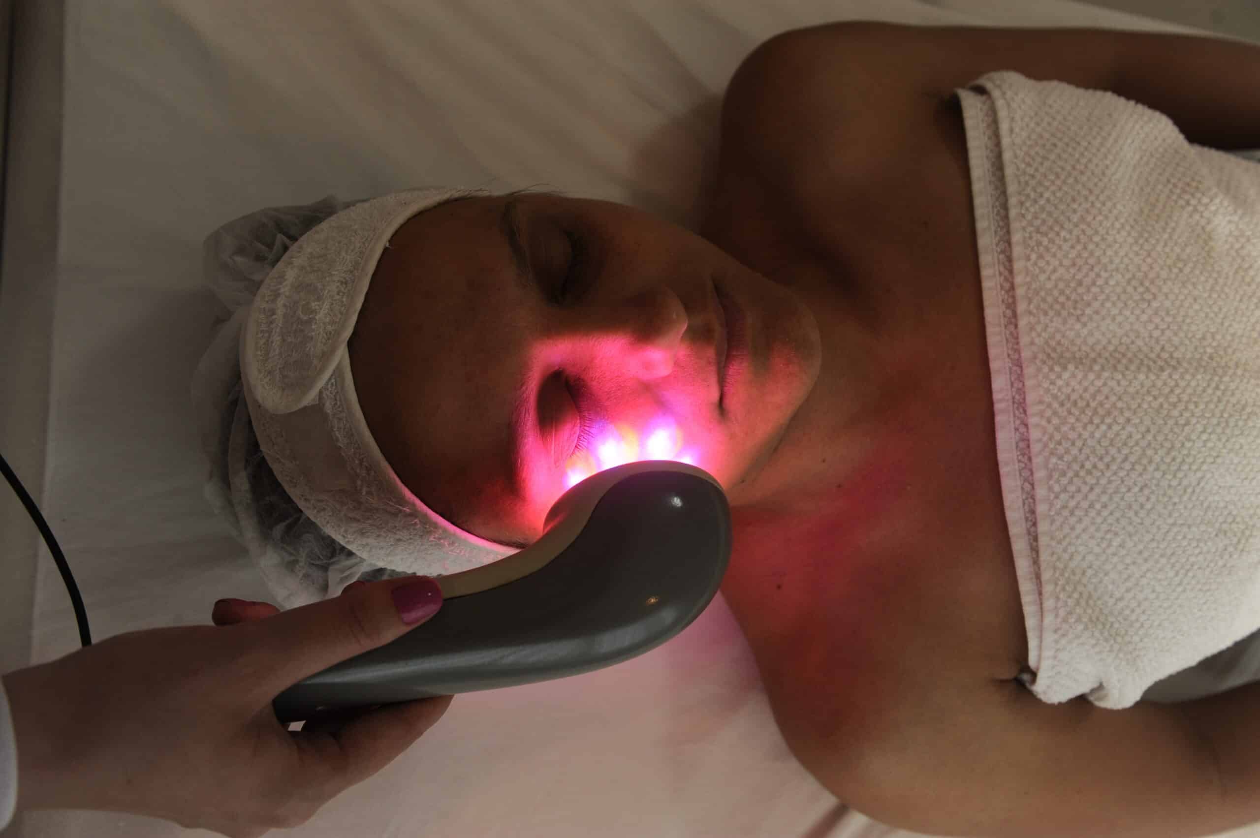 Femme reçoit un soin du visage par luminothérapie à lumière rouge. Une main tient l'appareil près de sa joue, elle porte un bandeau et une serviette.