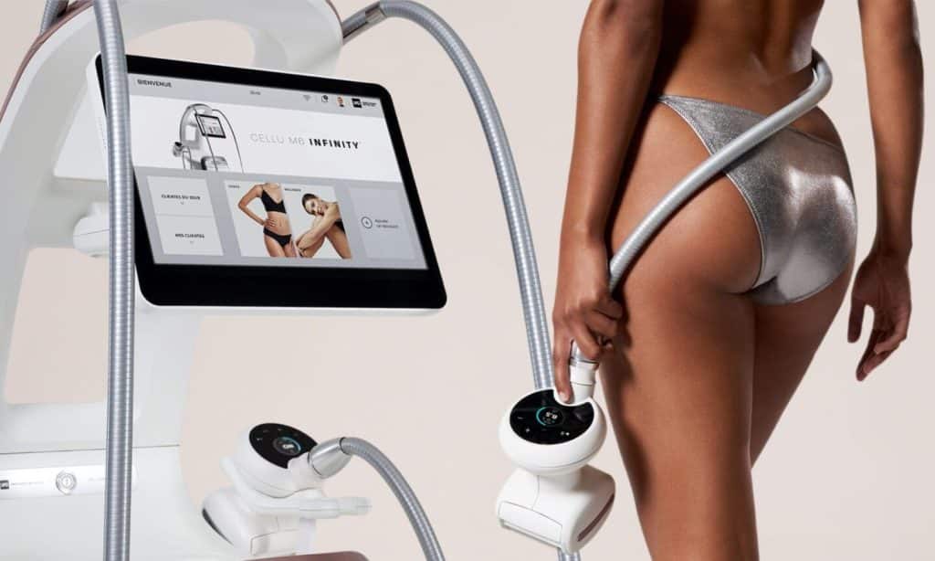 Femme en bikini argent tenant un applicateur de la machine CELLU MB INFINITY. L'écran affiche 'BIENVENUE' et des options de traitement corporel.