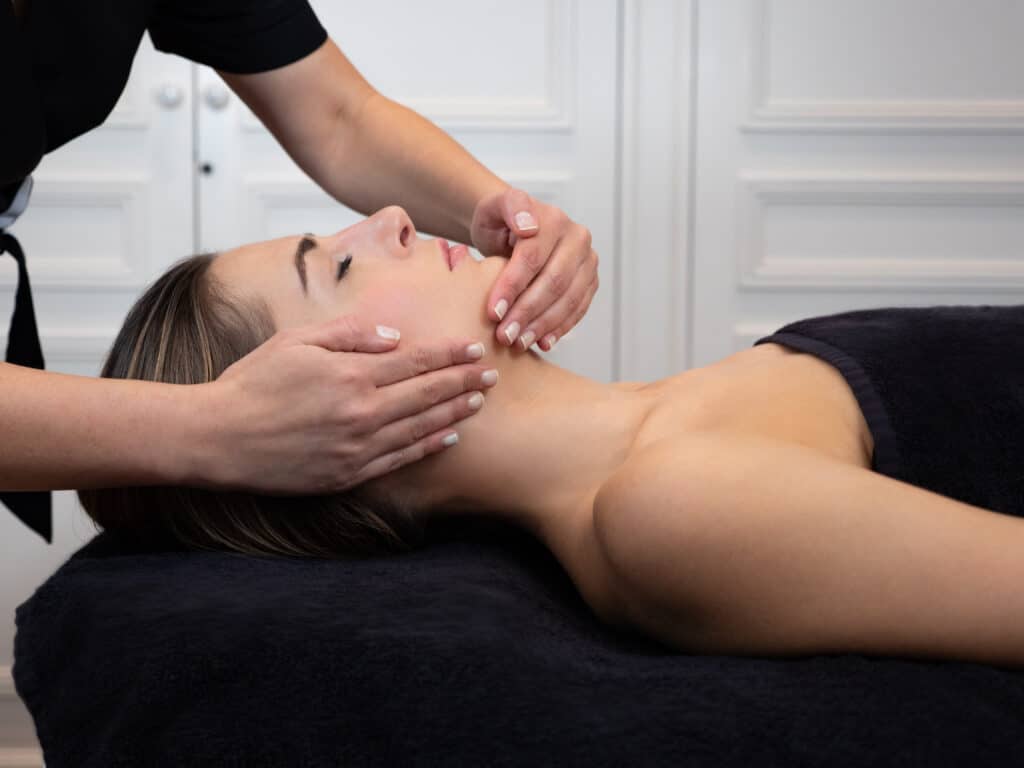 Femme recevant un massage facial relaxant par une esthéticienne, allongée sur une serviette noire dans un salon de beauté.